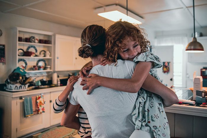 Mother carrying young daughters in home kitchen.