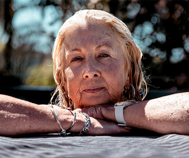 A mature woman with blonde hair leans on a pool edge wearing a watch and bracelets